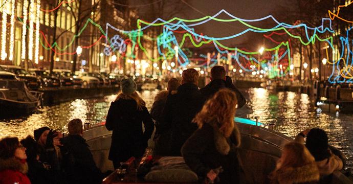 We see a boat with people approaching the artwork. The artwork is made up of coloured neon lines, forming figures above the canal. 